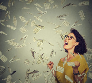 Portrait happy woman in glasses exults pumping fists ecstatic celebrates success under a money rain falling down dollar bills banknotes isolated on gray wall background with copy space
