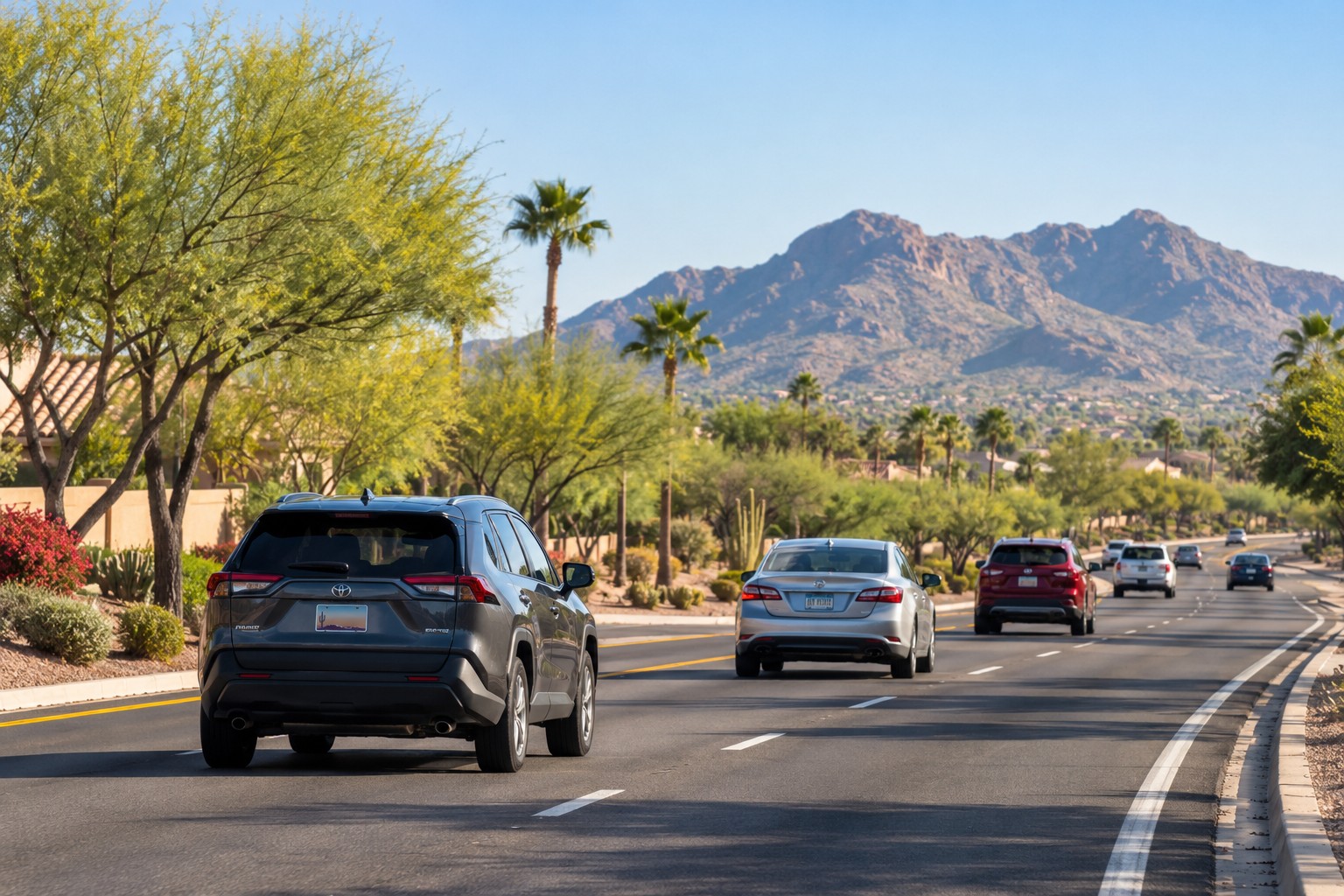 Vehicles with different license plates travel along a sunny Arizona boulevard lined with desert landscaping and palm trees, with mountains in the background.